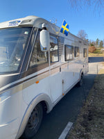 Svensk bilflagga monterad på husbil, visar flaggan sedd framifrån under en solig dag.