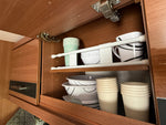 Vit teleskopstång i öppet träskåp med muggar, skålar och plastmuggar – smart kökförvaring i husbil och camping.